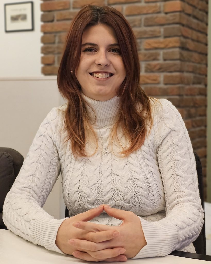 Shanees Pukies von NudgeMe Coaching sitzt lächelnd an einem Tisch in heller Umgebung mit weißem Pullover vor einer Backsteinwand.